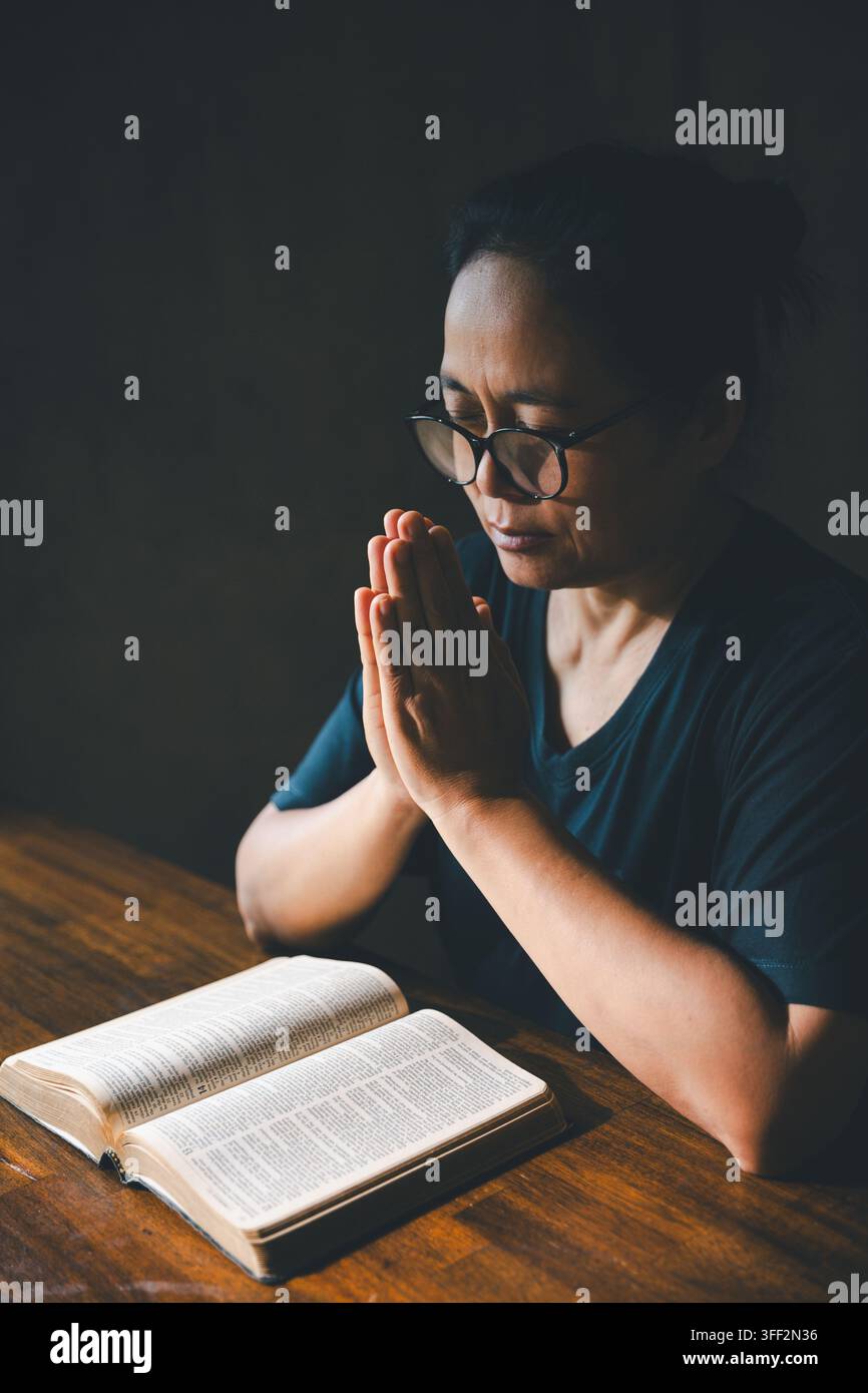 Person adulte portant des lunettes, priant les yeux fermés et les paumes jointes dans une pièce sombre, exprimant la foi, la dévotion, l'adoration, la spiritualité, l'espoir, la paix Banque D'Images