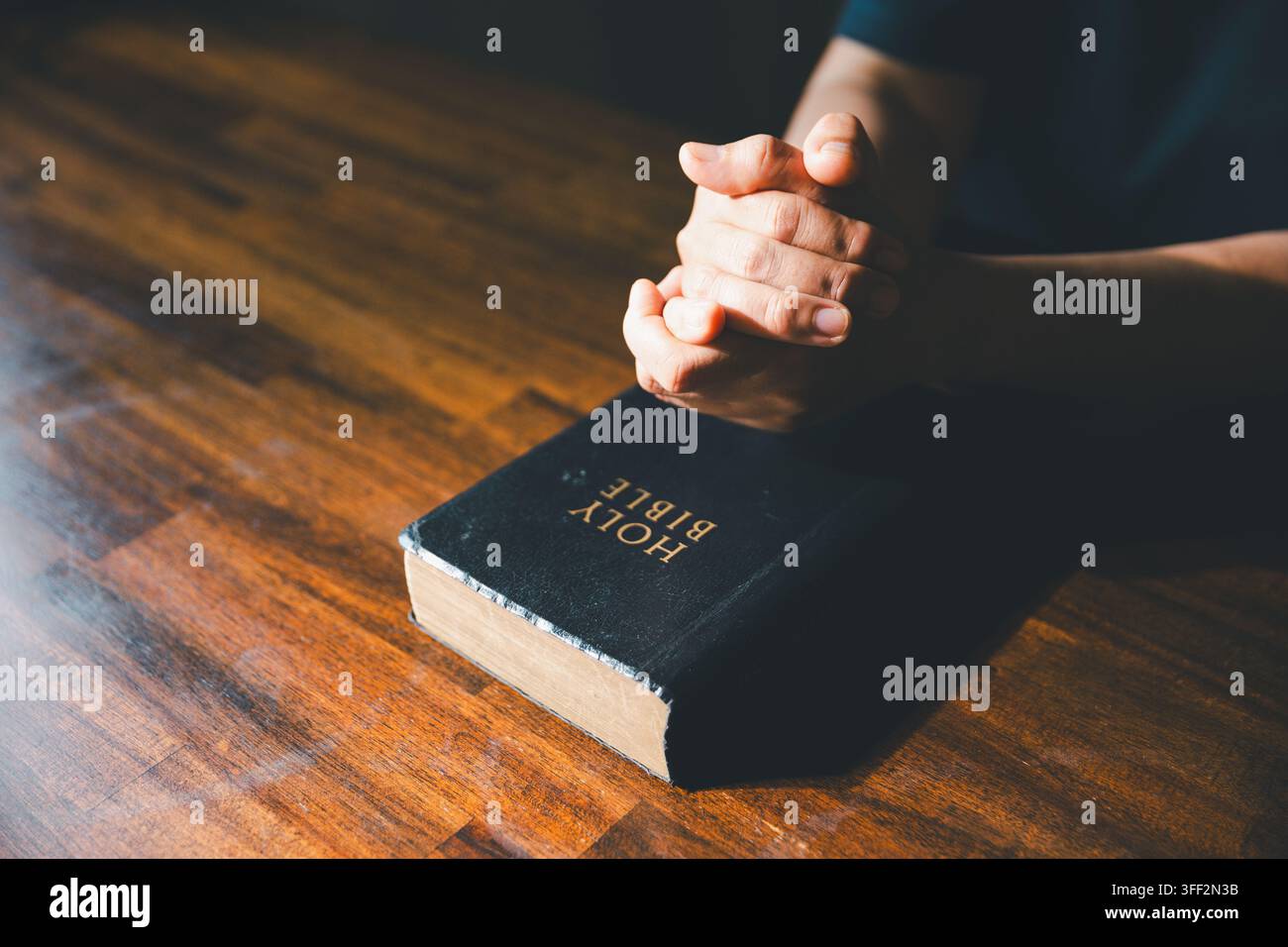 Gros plan de mains jointes au-dessus d'une Bible fermée sur une table en bois, symbolisant la prière, la foi chrétienne, la dévotion, l'adoration, les Écritures, le renouveau spirituel Banque D'Images