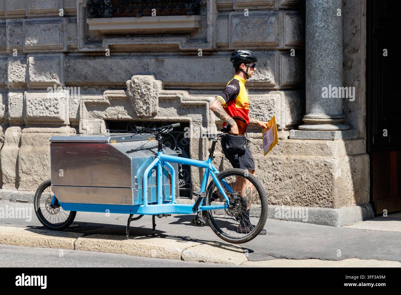 Courrier en uniforme DHL avec vélo cargo bleu et colis devant un bâtiment du centre-ville de Milan, Italie Banque D'Images