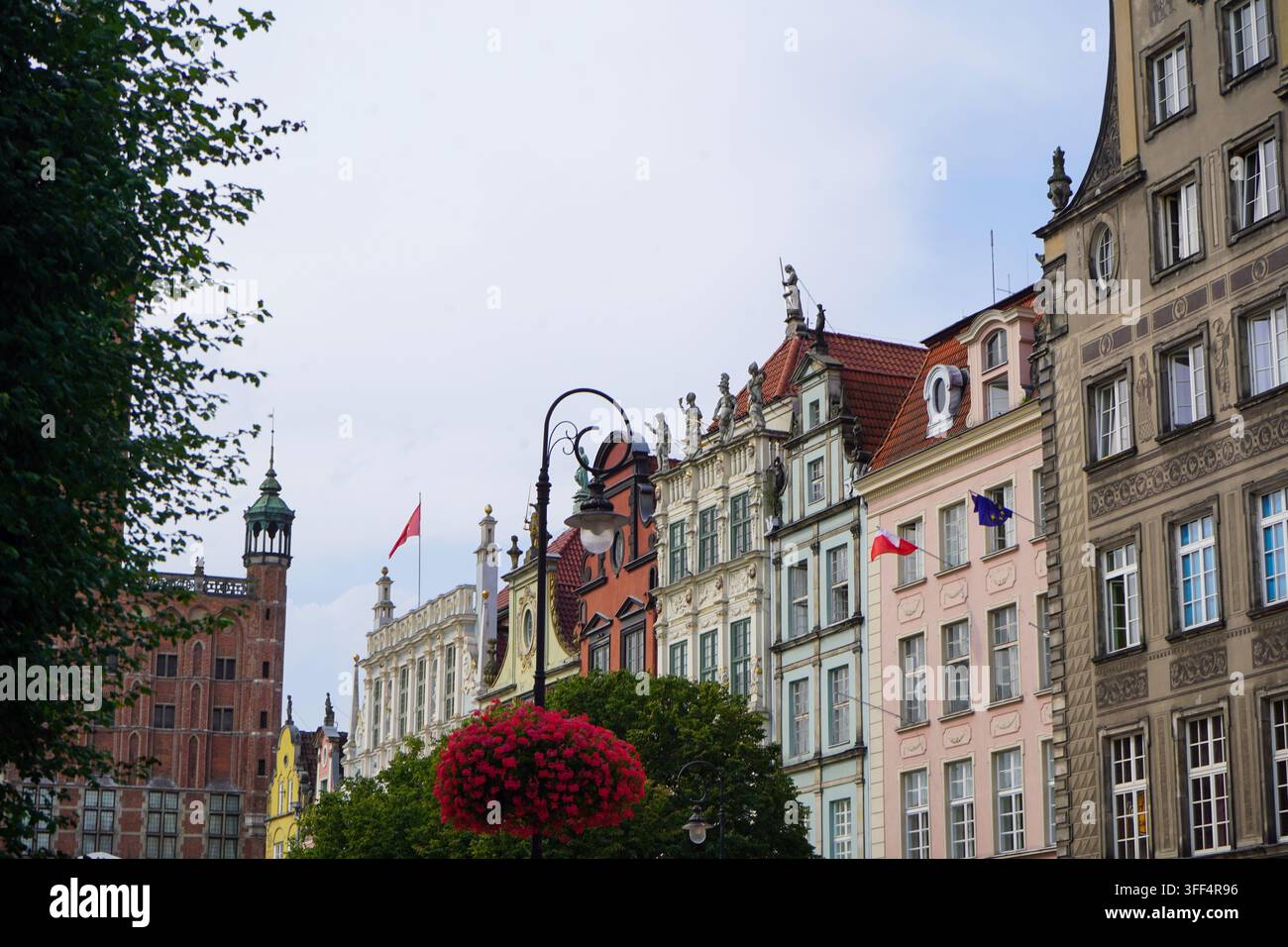 Des façades ornées élégantes et des toits traditionnels bordent une rue historique encadrée par des fleurs rouges suspendues et des arbres. Banque D'Images
