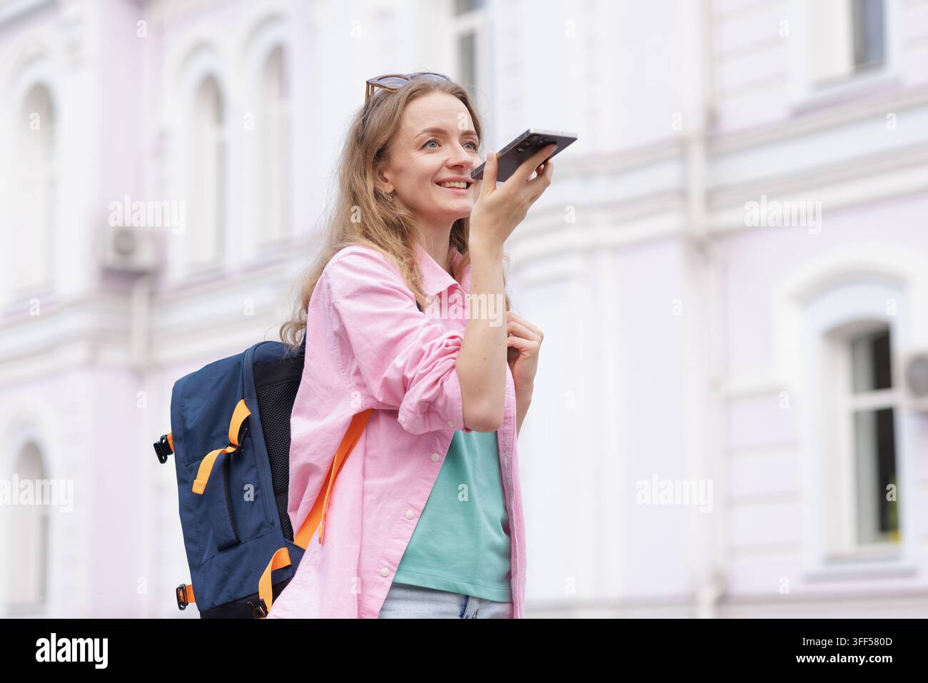 Jeune femme avec sac à dos utilise une assistante vocale sur smartphone en marchant en ville, illustrant la navigation de voyage, la technologie de recherche vocale et l'urbain Banque D'Images