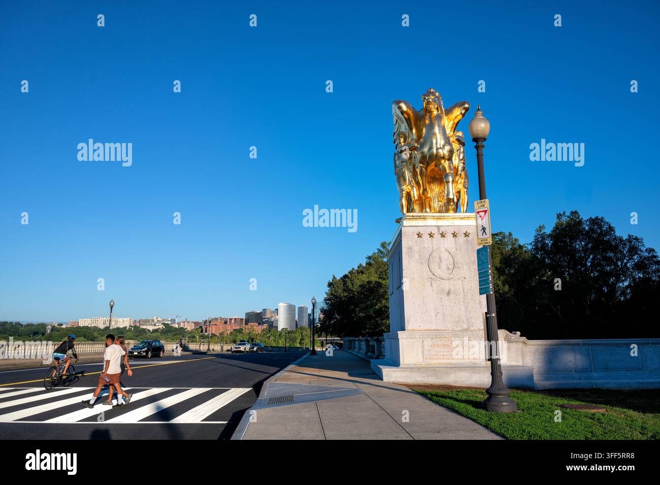 Statue « Aspiration and Literature » près du Lincoln Memorial | WASHINGTON, D.C. — Des piétons et un cycliste passent devant « Aspiration and Literature » sur Parkway Drive NW près du Lincoln Memorial, dimanche 6 septembre 2026. James Earle Fraser a sculpté le groupe équestre en bronze doré, coulé à Rome en 1950 en cadeau de l’Italie aux États-Unis. Il est dressé sur un piédestal en granit marquant l’entrée du Rock Creek and Potomac Parkway près du pont commémoratif d’Arlington. La ligne d’horizon de Rosslyn, en Virginie, s’élève de l’autre côté du fleuve Potomac en arrière-plan. Banque D'Images