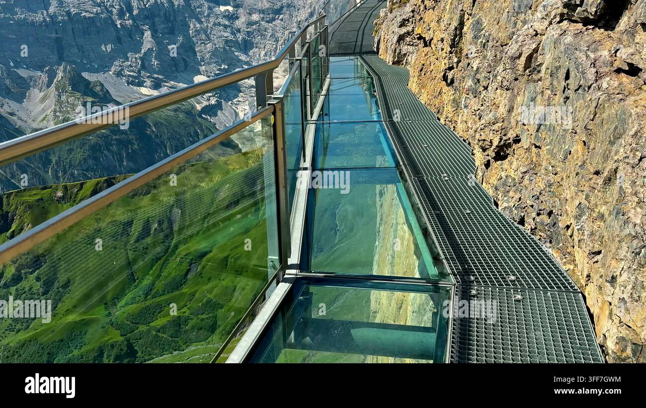 Passage vitrée au-dessus d'une vallée alpine dramatique. La passerelle d'observation en verre épouse une falaise rocheuse escarpée au-dessus des pentes alpines verdoyantes et des montagnes escarpées Banque D'Images