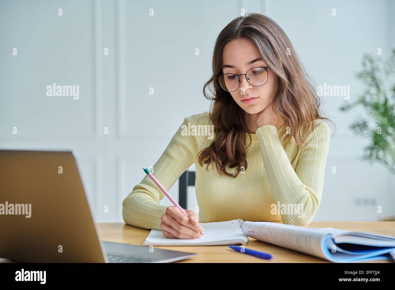 Étudiante sérieuse portant des lunettes, écrivant attentivement dans un cahier devant un ordinateur portable. Concept de fatigue d'étude, apprentissage à distance intensif, Banque D'Images