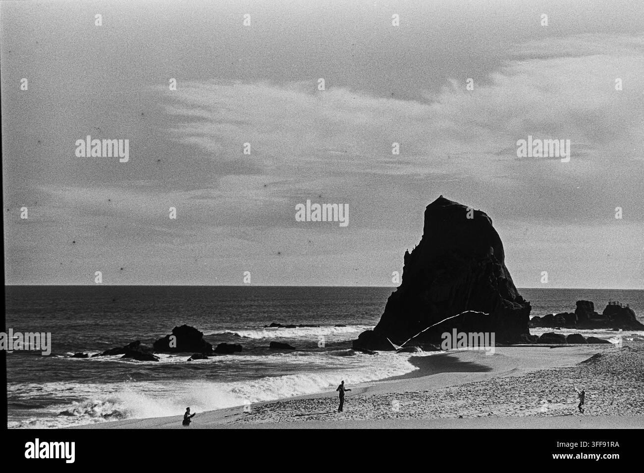 Une tour rocheuse sombre domine une plage de sable avec trois personnes au loin dans une scène de film analogique noir et blanc granuleux. Banque D'Images