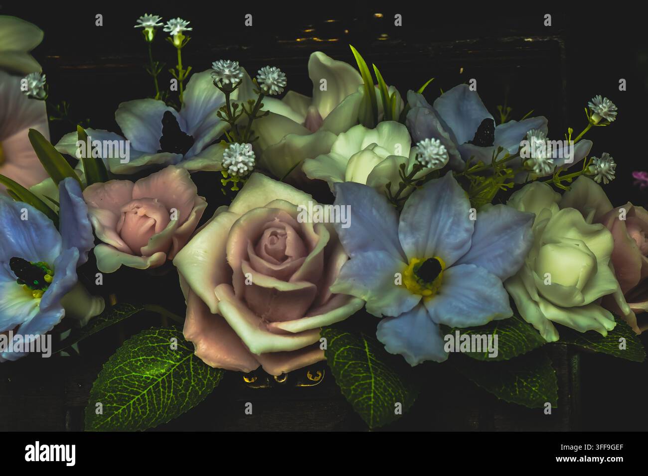 Roses artificielles et fleurs bleues arrangées avec de petites fleurs blanches et des feuilles vertes sur un fond sombre pour une décoration florale élégante. Banque D'Images