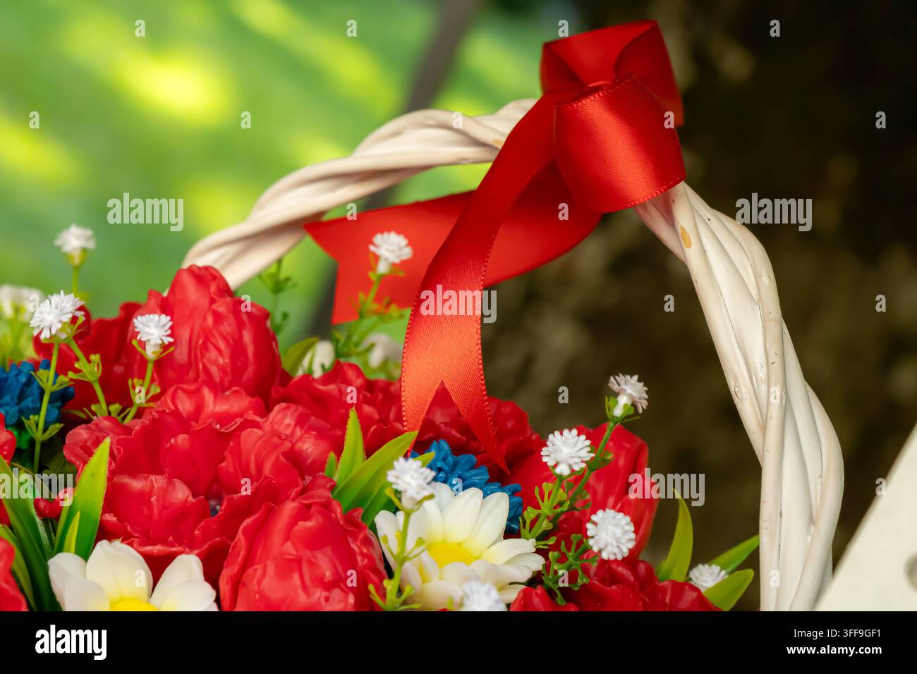 Fleurs artificielles rouges, marguerites blanches et feuilles vertes disposées dans un panier en osier blanc avec un nœud en satin, créant une décoration florale lumineuse Banque D'Images