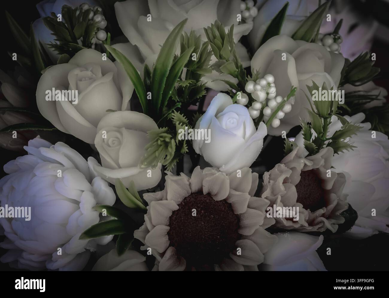 Gros plan de roses blanches artificielles, d'accents de tournesol et de feuillage vert disposés sur un fond sombre pour une décoration florale élégante. Banque D'Images