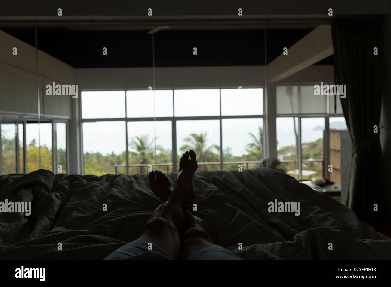 Une personne se repose sur un lit défait dans une chambre sombre, les pieds levés et en silhouette devant de grandes fenêtres panoramiques révélant des palmiers tropicaux luxuriants et Banque D'Images