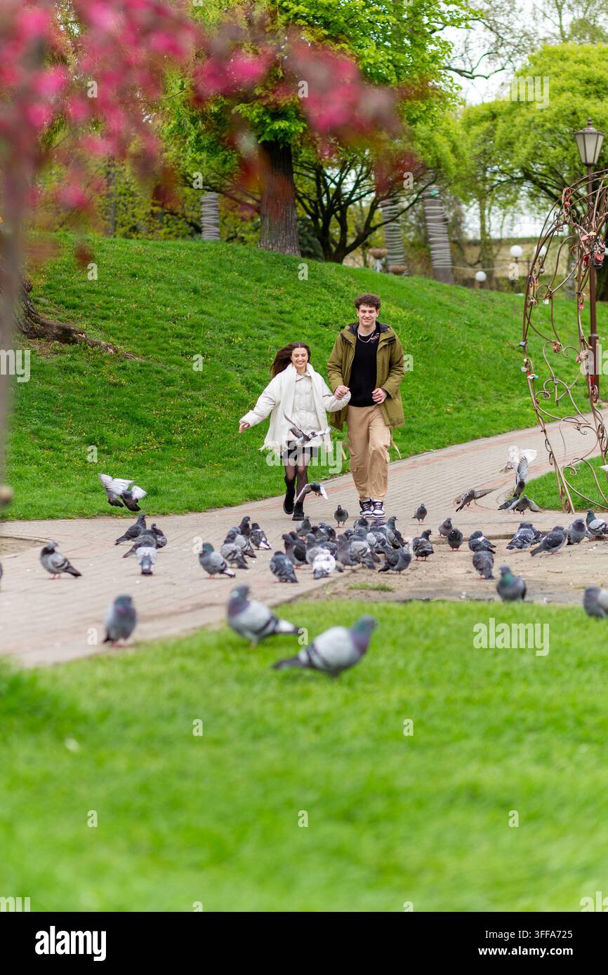 Un jeune couple heureux se tenant par la main et courant le long d'un sentier du parc tandis que des pigeons volent autour d'eux au printemps. Banque D'Images