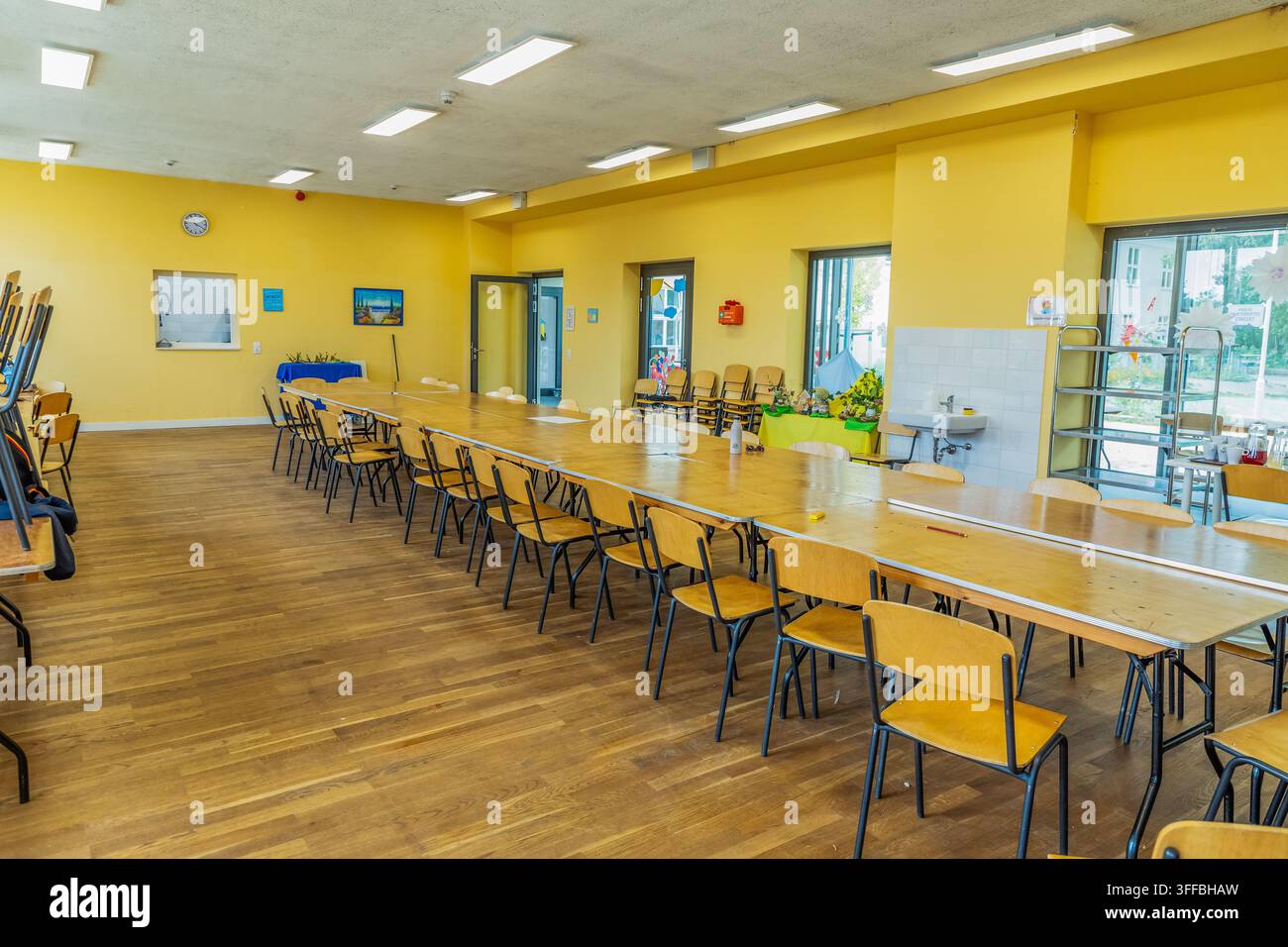 Une salle à manger commune lumineuse présente une longue table en bois avec des chaises disposées, créant une atmosphère accueillante pour les rassemblements et les repas. Banque D'Images