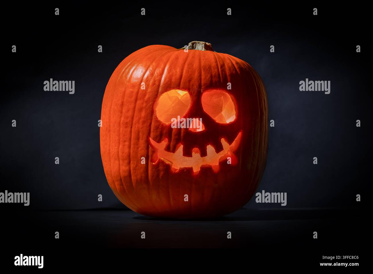 Jack-o'-lantern orange illuminé avec une tête sculptée ressemblant à un crâne, présentant des yeux ronds et des dents déchiquetées, éclairé de l'intérieur sur un fond sombre dramatique en studio Banque D'Images