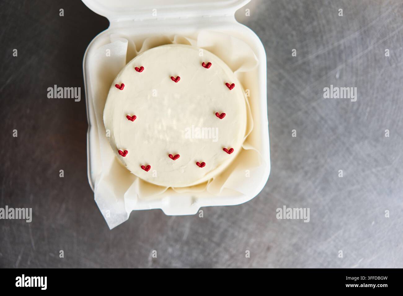 Gâteau rond blanc givré avec de petites décorations en forme de cœur rouge dans une boîte à emporter sur une surface métallique, avec gâteau, glaçage, blanc, détail en cœur, montrant le rouge Banque D'Images