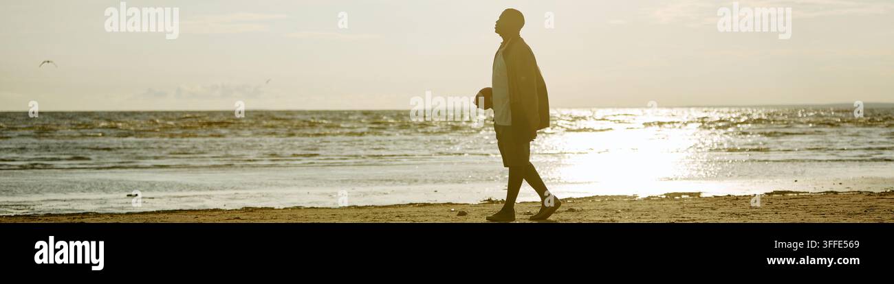Tête d'un homme noir marchant sur le rivage de la plage tenant un ballon de football au coucher du soleil Banque D'Images