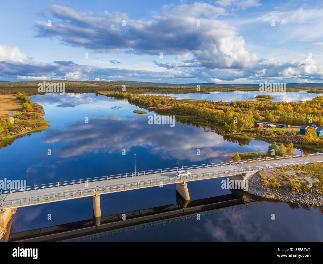 Vue aérienne d'une voiture blanche traversant un pont moderne au-dessus d'un lac miroir reflétant un ciel nuageux dramatique dans la Laponie arctique, en Suède Banque D'Images