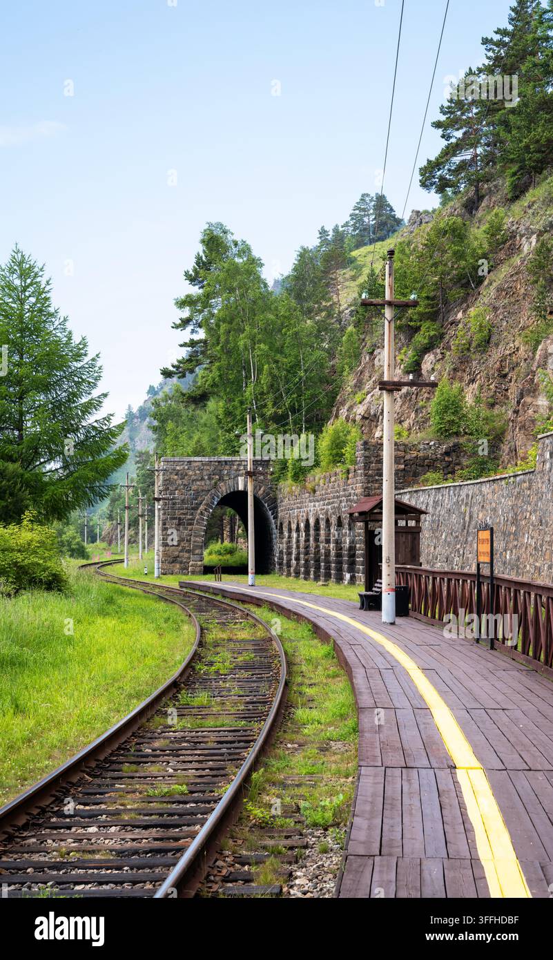 Une photographie verticale montrant une voie ferrée à voie unique incurvée menant à un tunnel en arche de pierre sombre construit dans une falaise rocheuse. À droite du t Banque D'Images
