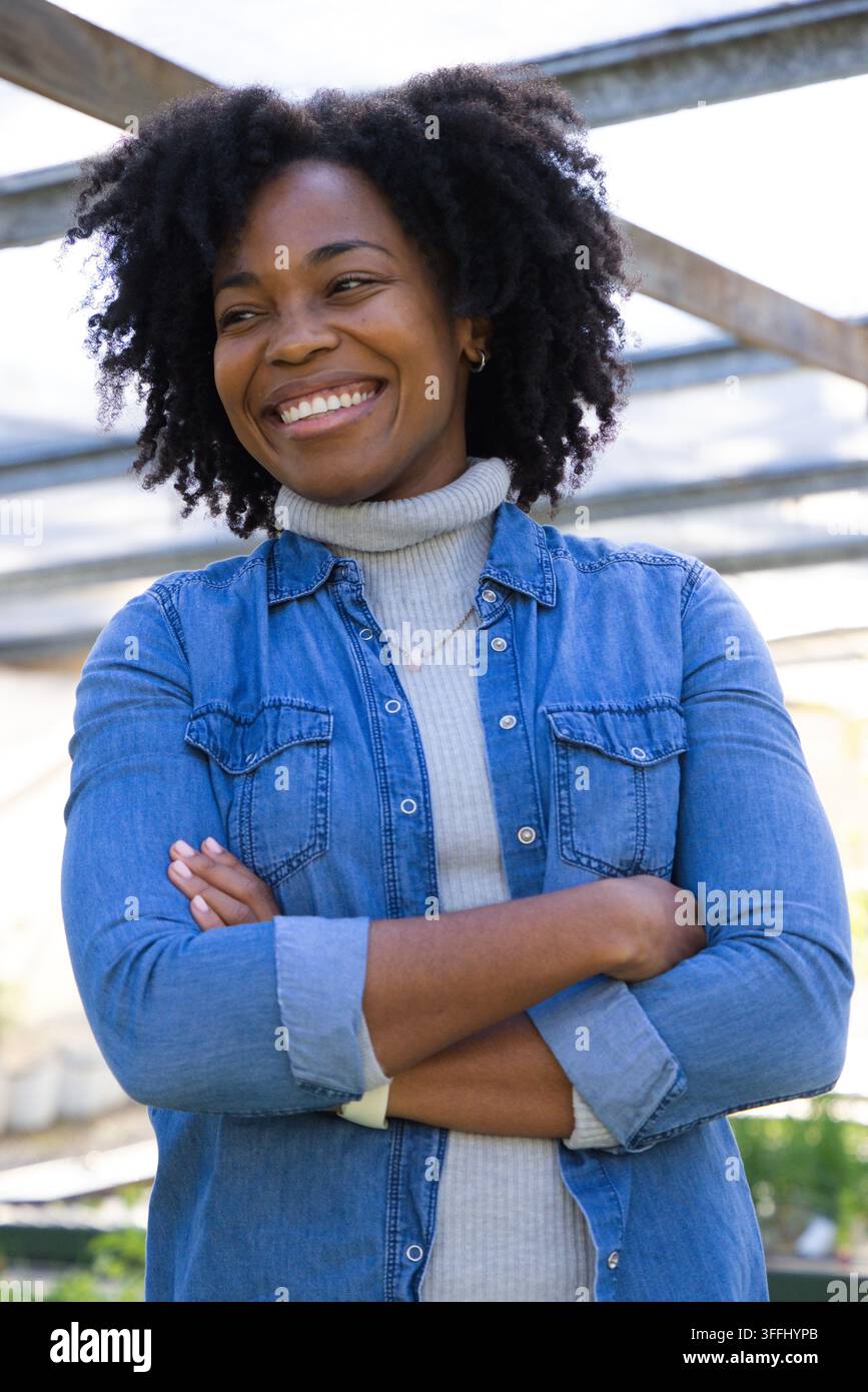 Femme afro-américaine d'âge moyen debout sous une pergola métallique, souriante, portant une chemise en jean et un collier Banque D'Images
