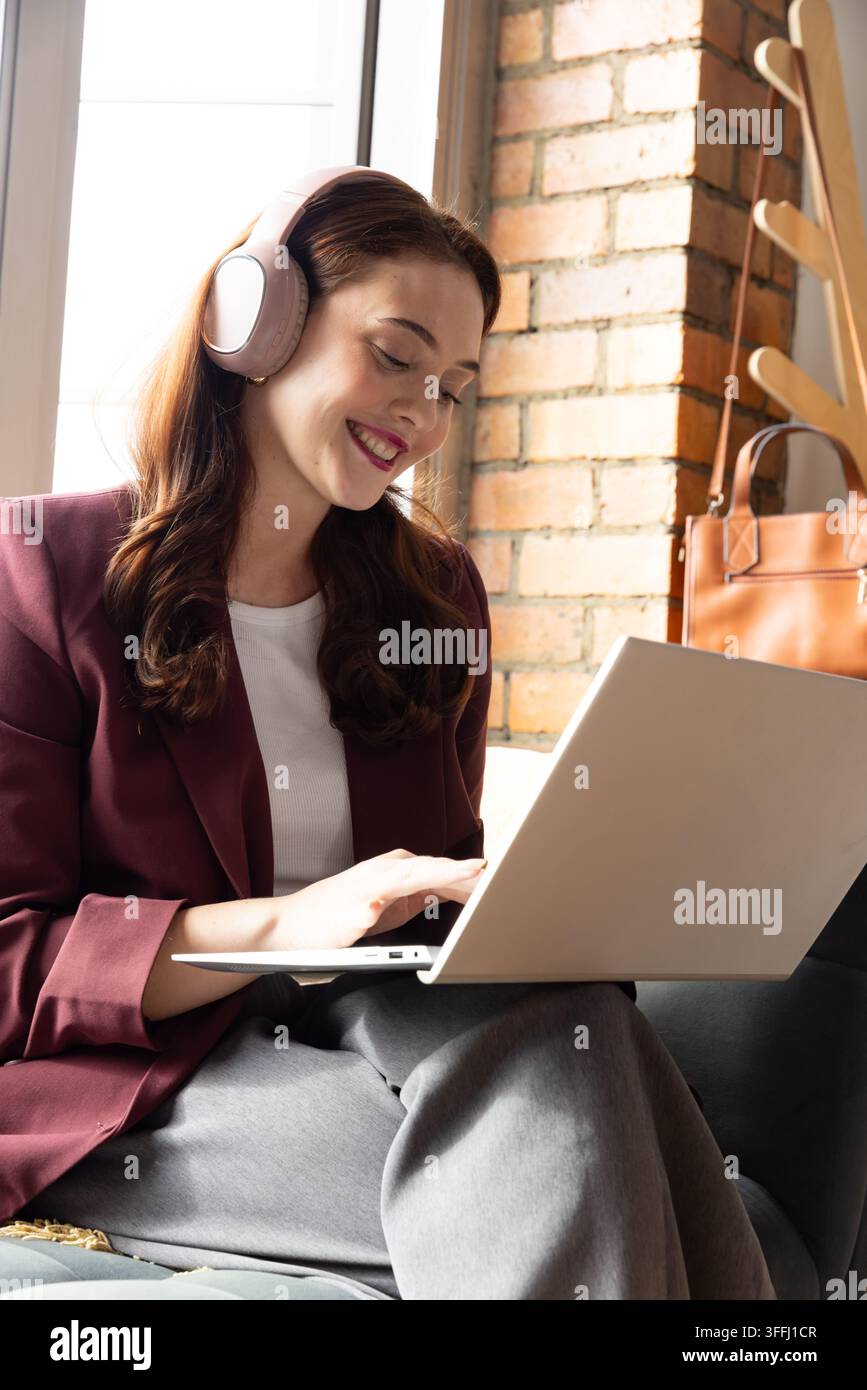 Femme assise sur un canapé près de la fenêtre, portant un blazer bordeaux et un casque audio, utilisant un ordinateur portable Banque D'Images