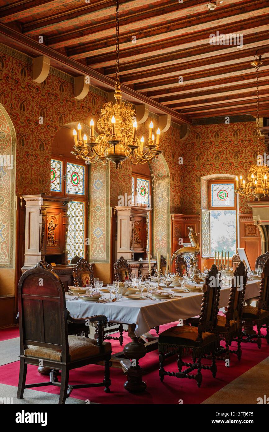Salle à manger d'un château médiéval opulent avec des murs tapissés de riches tapisseries, des meubles en chêne sculptés d'époque, des vitraux et de grands lustres Banque D'Images