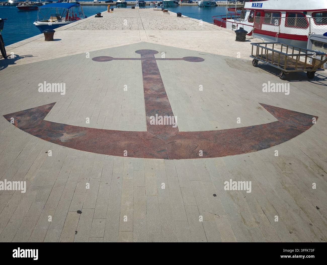 Une mosaïque d'ancre en pierre rouge détaillée incrustée dans le quai du port, entourée de bateaux dans une marina côtière méditerranéenne sereine. - Image de stock capturée avec un smartphone