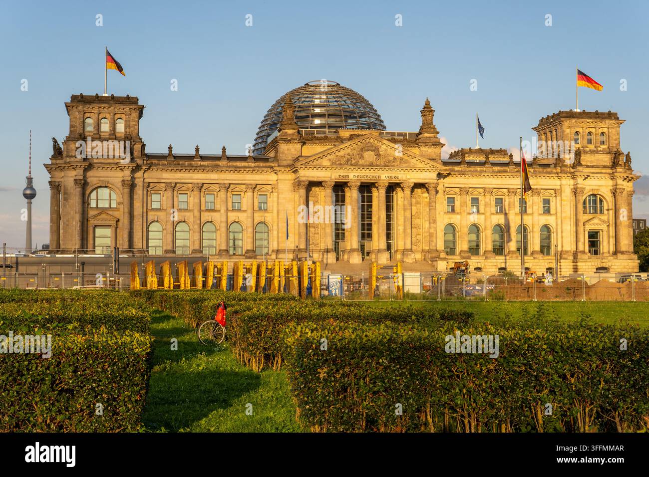 Berlin, Allemagne - 01 août 2026 : Une personne portant un sac rouge et un vélo marche à travers des haies taillées devant un grand bâtiment en pierre avec une porte en verre Banque D'Images