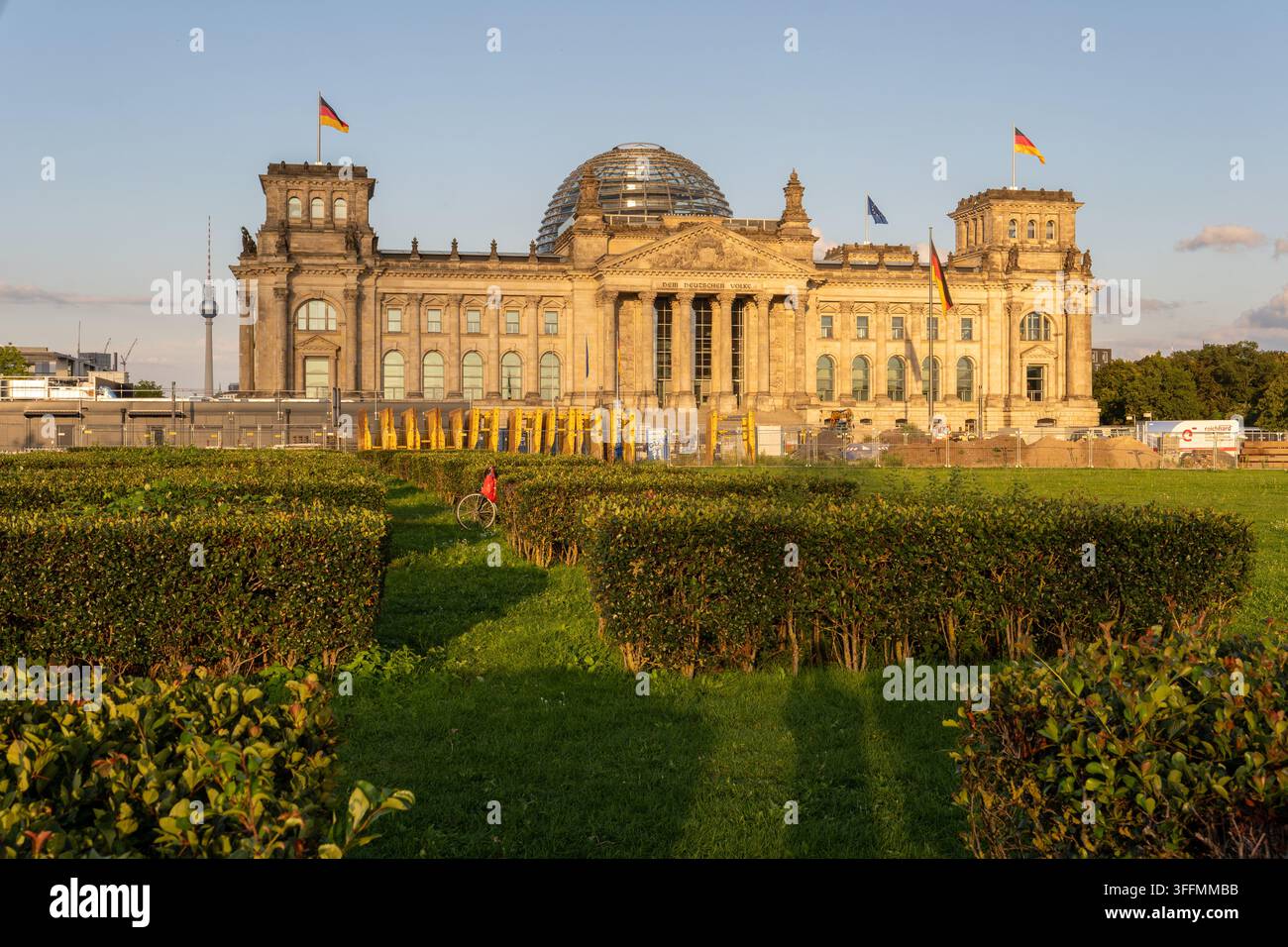Berlin, Allemagne - 1er août 2026 : Une personne en veste rouge fait du vélo le long d'un chemin herbeux bordé de haies taillées devant un bâtiment en pierre Banque D'Images