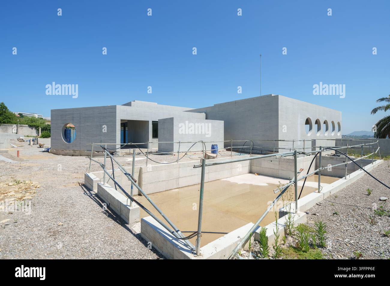 Bâtiment d'hôtel contemporain en construction sur un chantier graveleux ensoleillé, avec des murs en béton inachevés, des ouvertures de fenêtres circulaires et une piscine encastrée Banque D'Images