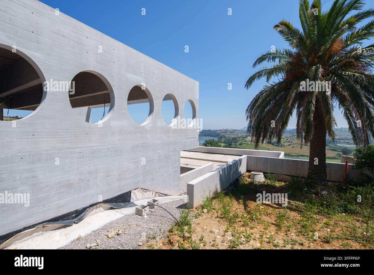 Hôtel contemporain en construction avec des ouvertures de fenêtres circulaires, des murs en béton apparents et un palmier surplombant un paysage rural sous un cl Banque D'Images