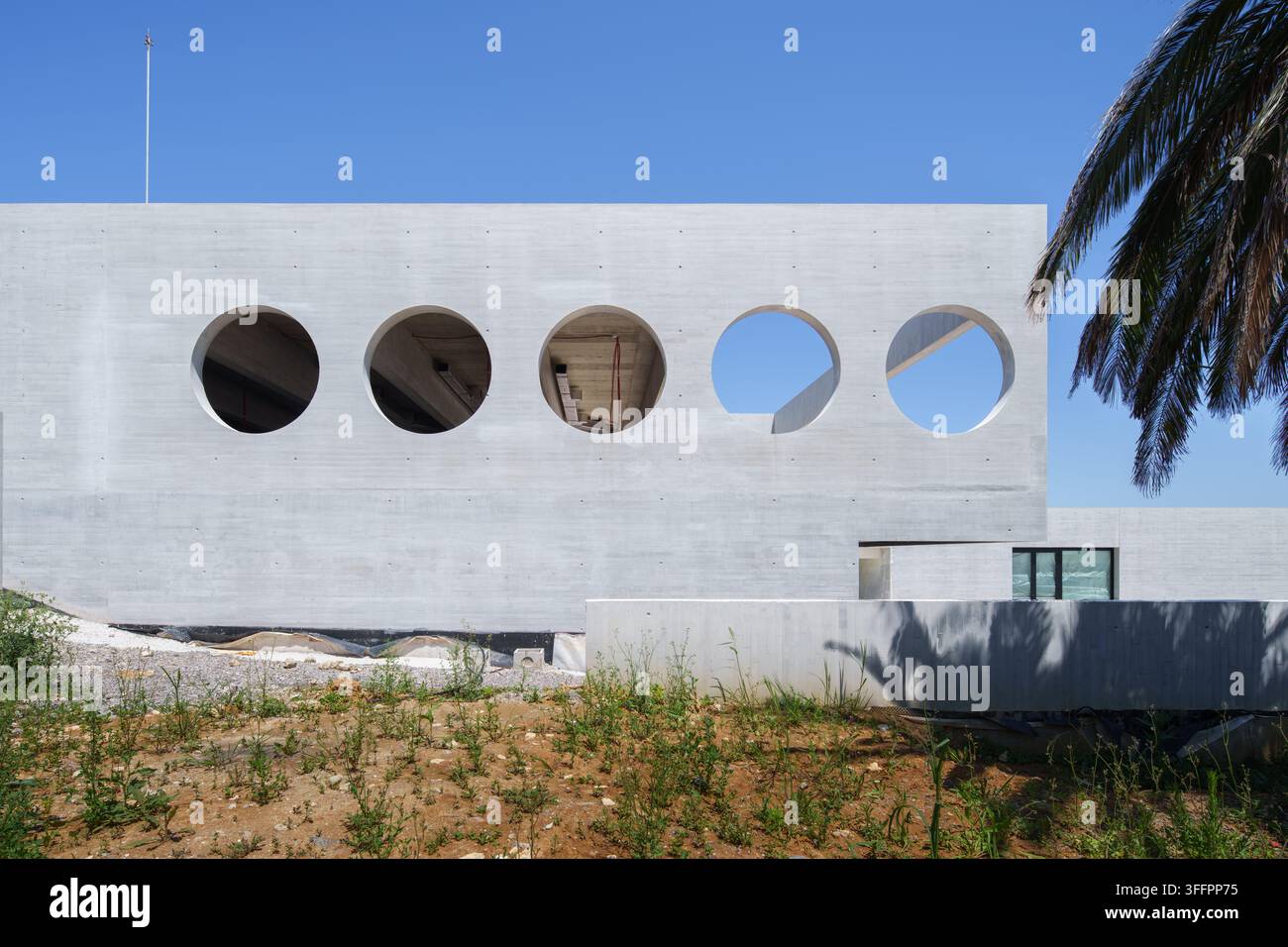 Bâtiment d'hôtel contemporain en béton blanc en construction, présentant une rangée d'ouvertures de fenêtres circulaires, un sol inachevé, l'ombre d'un palmier et c Banque D'Images
