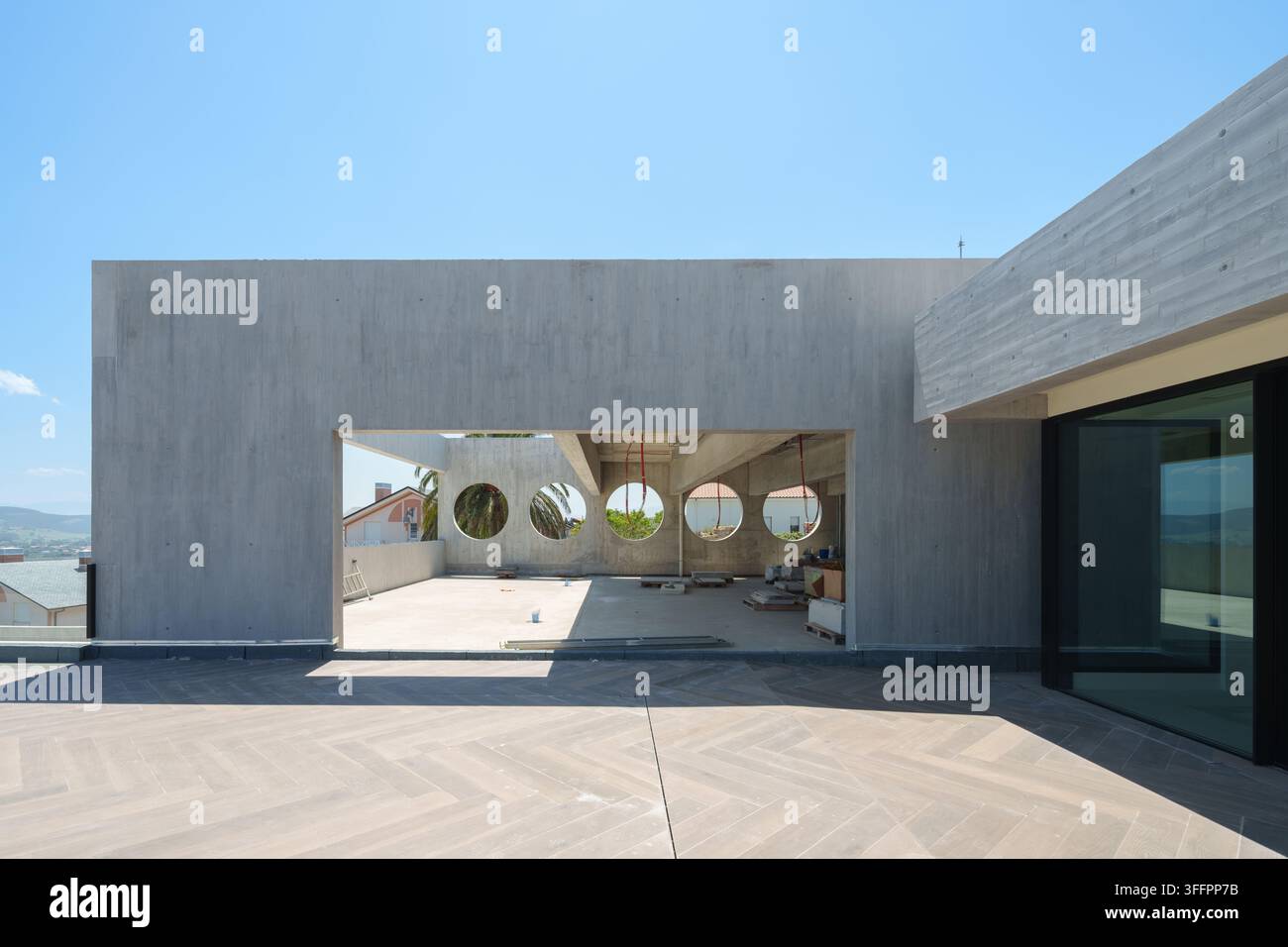 Construction hôtelière contemporaine présentant une façade en béton minimaliste, des ouvertures de fenêtres circulaires et une terrasse carrelée spacieuse sous un ciel bleu clair. Banque D'Images