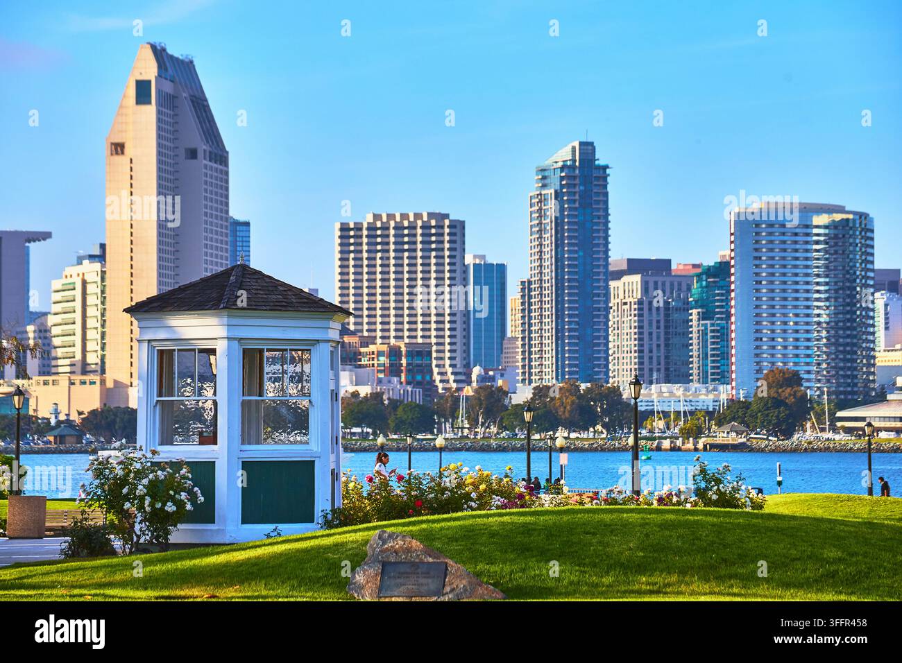La lumière du soleil baigne la skyline de San Diego alors qu'elle se dresse au-delà du parc de Coronado avec ses pelouses verdoyantes et son charmant pavillon blanc au bord de la baie. Des fleurs colorées Banque D'Images