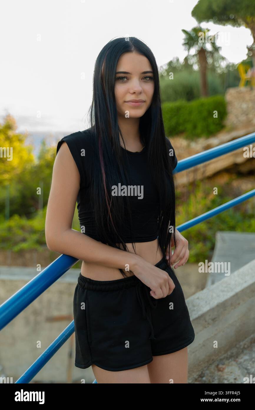 Jeune femme en tenue noire se tient à l'extérieur près de rampes bleues avec une expression calme. Banque D'Images