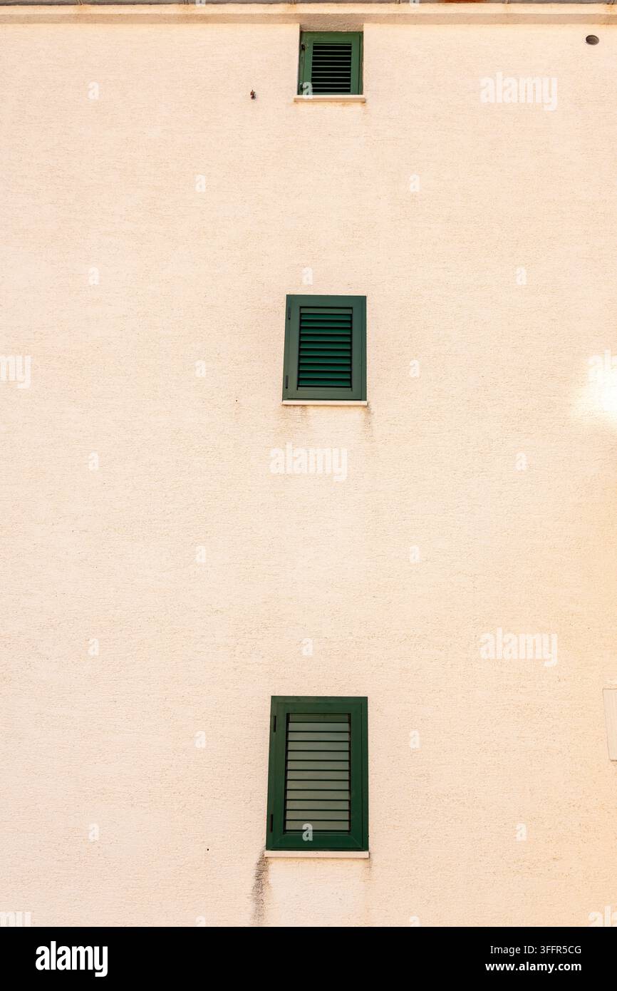 Une façade de bâtiment beige sobre présente trois petites fenêtres à volets verts disposées verticalement. Banque D'Images