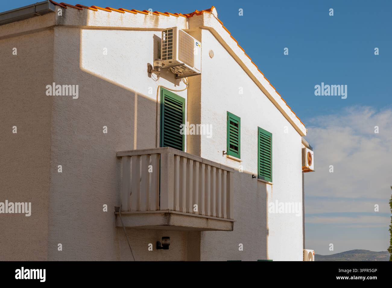 Une maison en stuc blanc avec des volets verts, un petit balcon et des climatiseurs muraux sous un ciel bleu. Banque D'Images