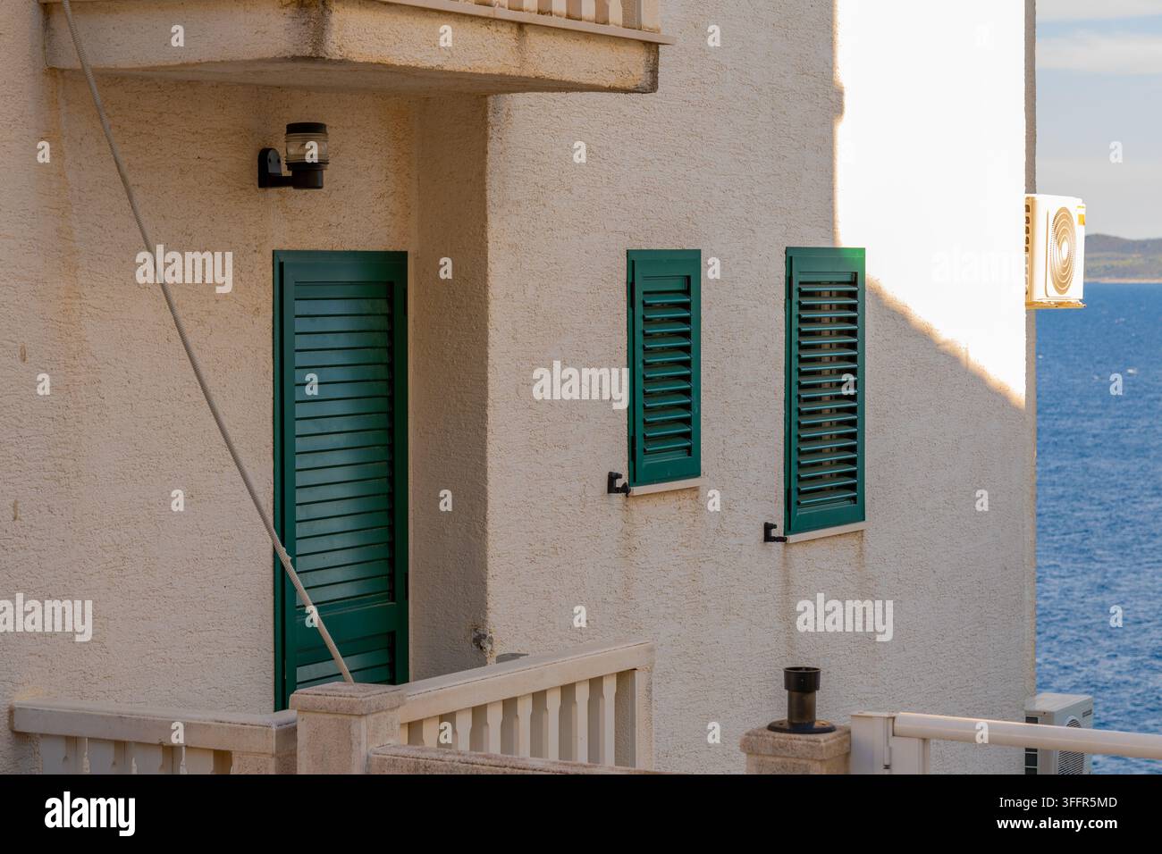 Une façade de maison ensoleillée avec des volets verts se dresse au bord de la mer bleue. Banque D'Images
