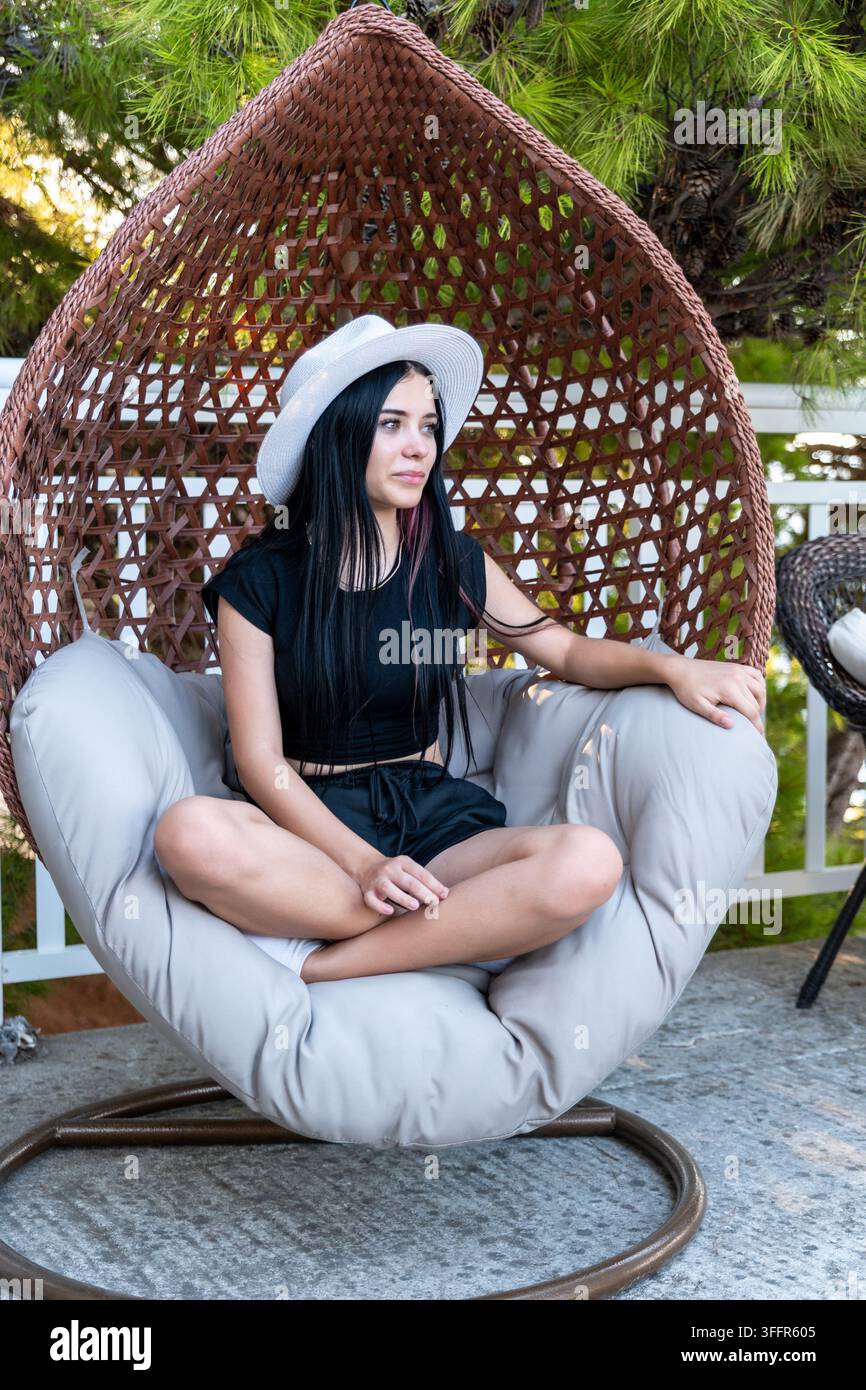 Jeune femme portant un chapeau blanc est assise en tailleur dans un fauteuil suspendu tissé en plein air. Banque D'Images