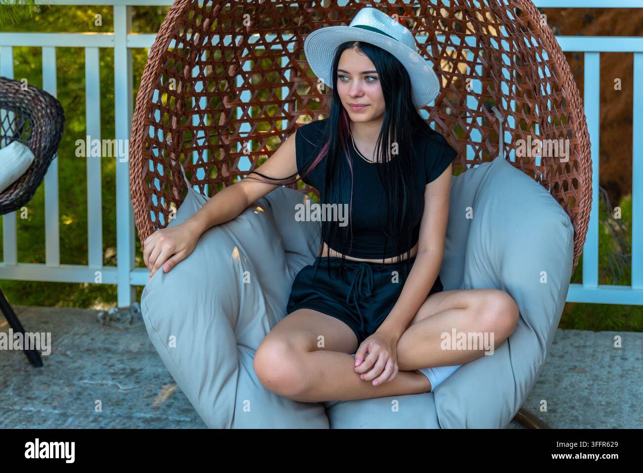 Une jeune femme est assise en tailleur dans une chaise suspendue tissée en plein air, portant un chapeau léger. Banque D'Images