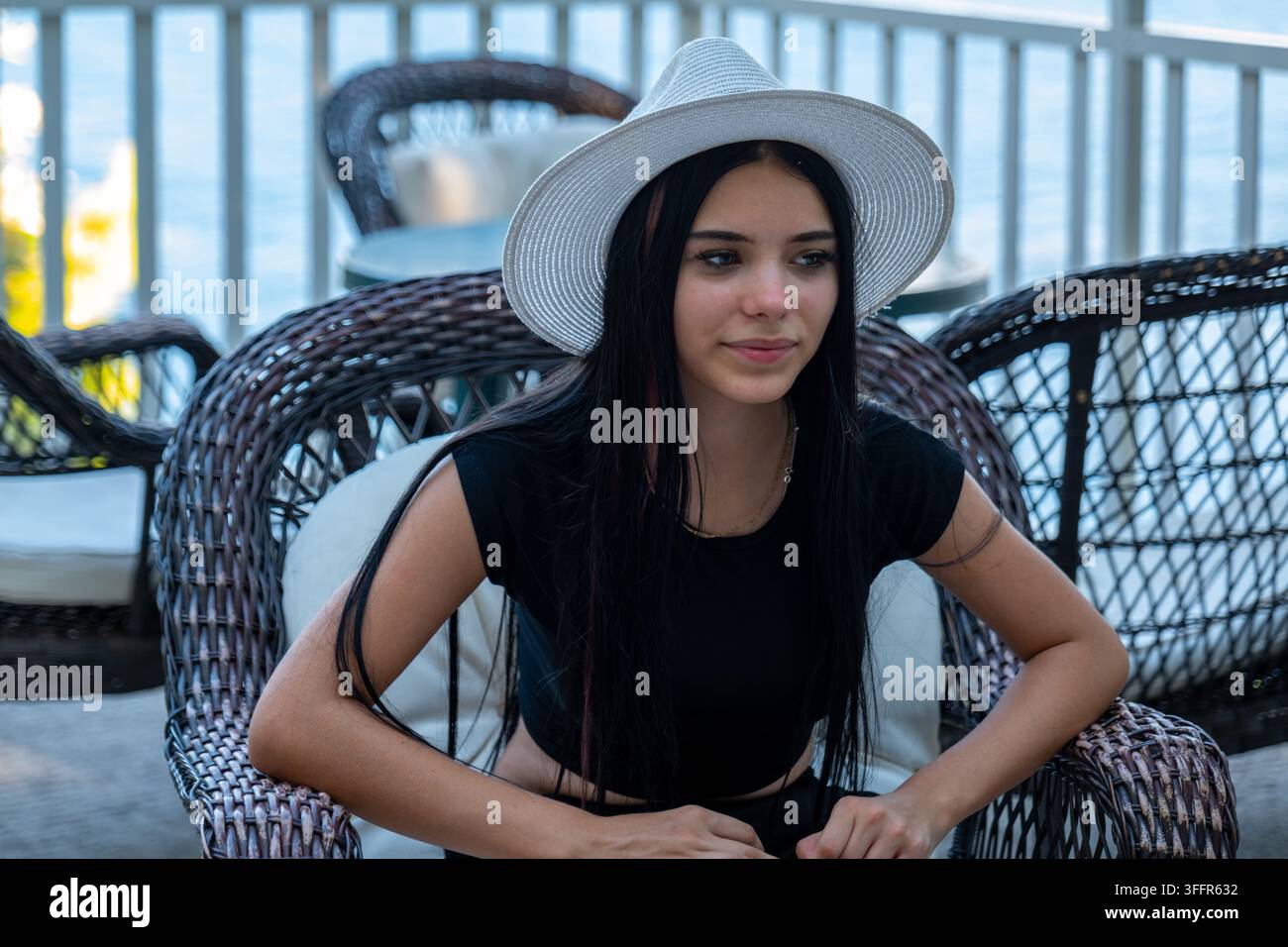 Jeune femme portant un chapeau blanc assise sur une chaise en osier près de l'eau. Banque D'Images