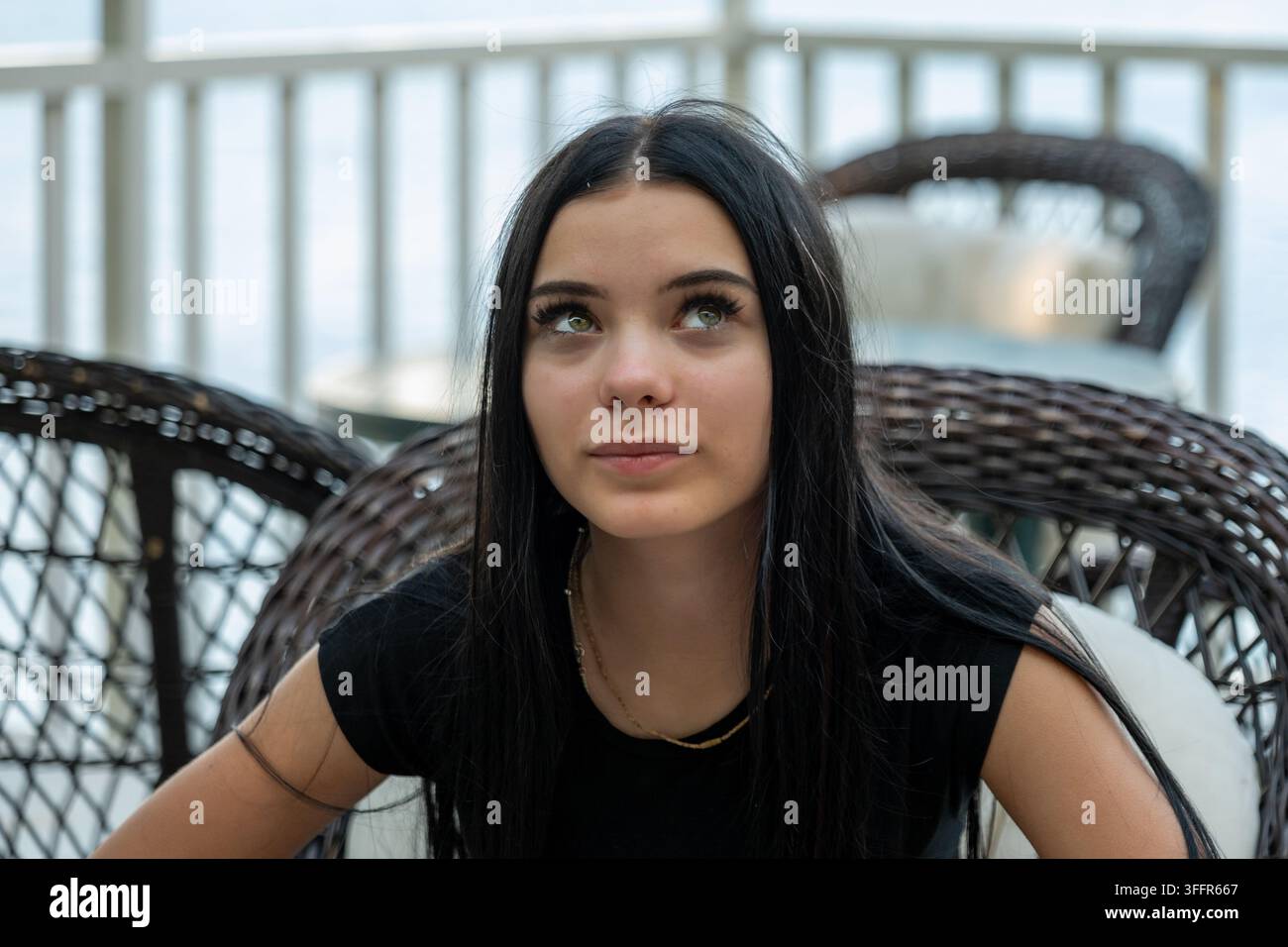 Une jeune femme aux longs cheveux foncés est assise dans une chaise en osier et regarde pensivement vers le haut. Banque D'Images