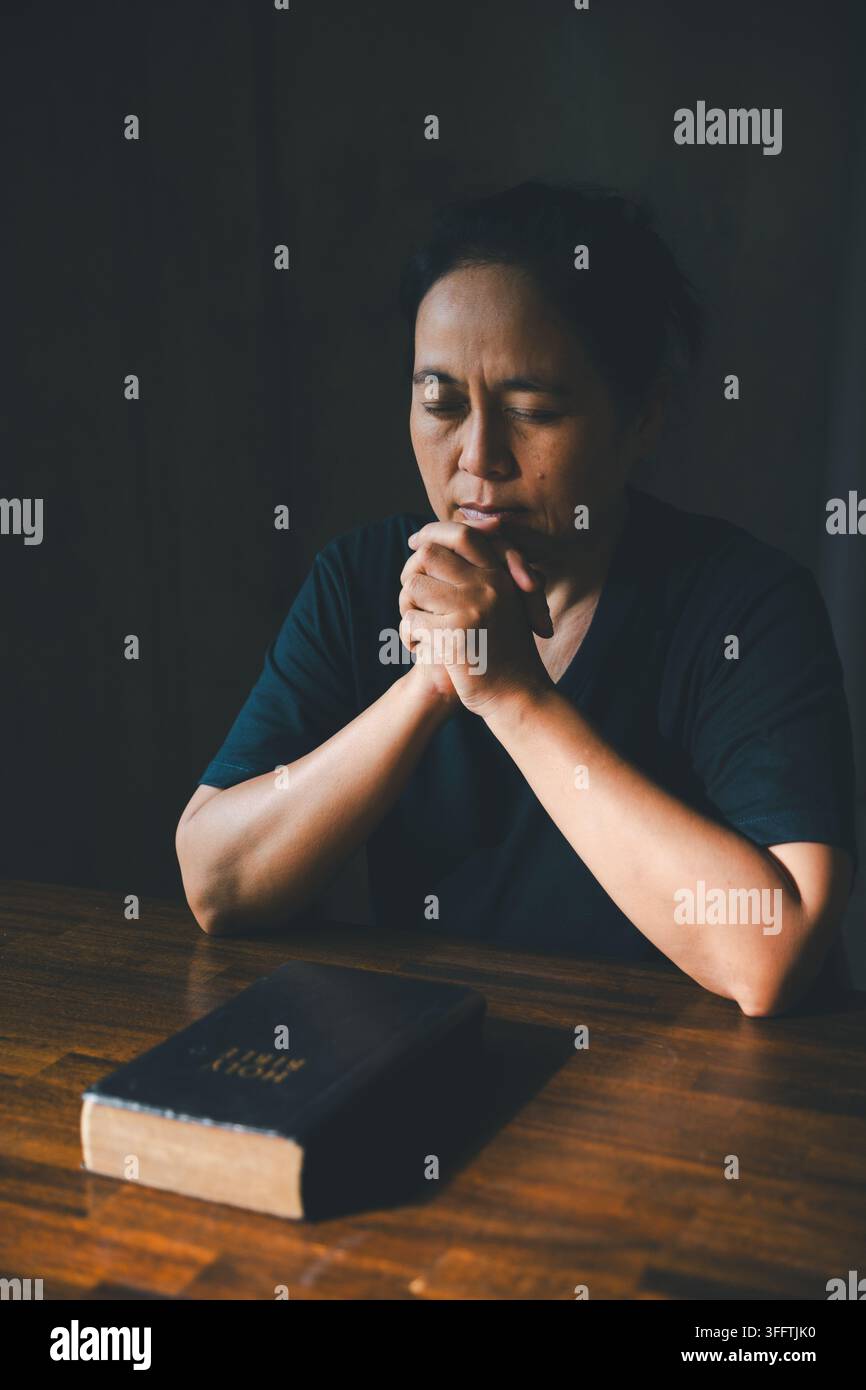 Personne adulte priant les mains jointes et la tête inclinée sur une Bible posée sur une table en bois dans une pièce sombre, exprimant la foi chrétienne, la dévotion, l'adoration, h Banque D'Images