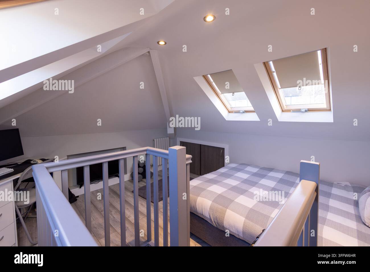 Chambre mansardée convertie vue à travers une balustrade d'escalier peinte avec fenêtres de toit pour lit double, plafonds inclinés, éclairages encastrés, bureau, télévision, rangement Banque D'Images
