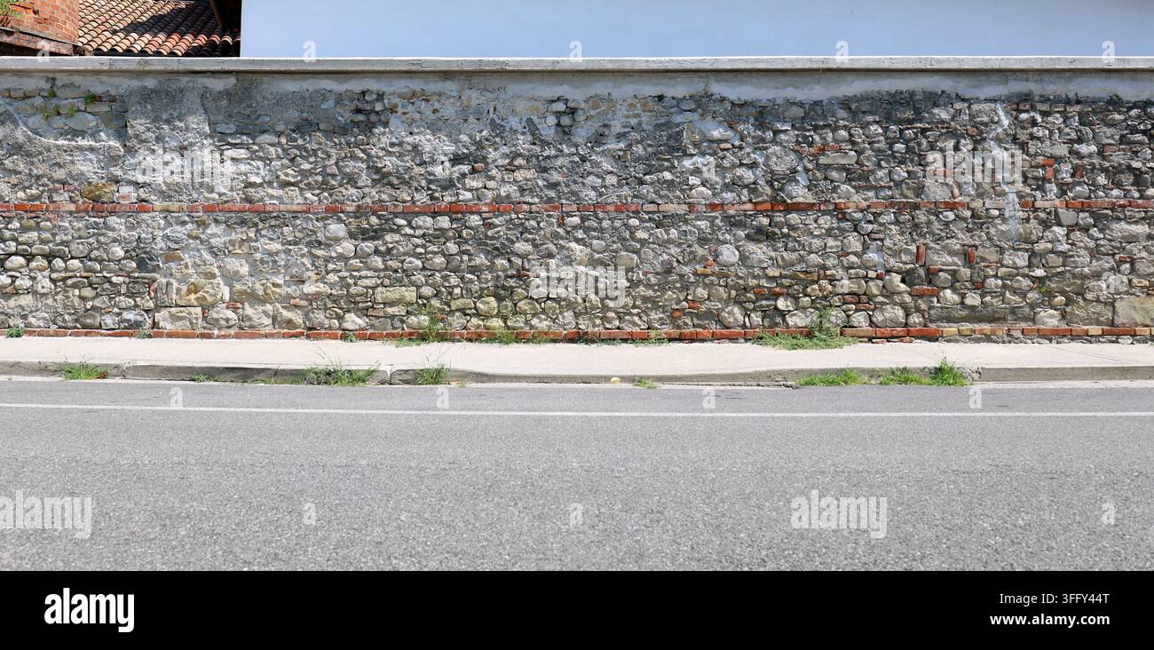 Long mur de pierre entourant fait de roches naturelles et rang de briques, trottoir en béton et route devant. Fond pour espace de copie. Banque D'Images