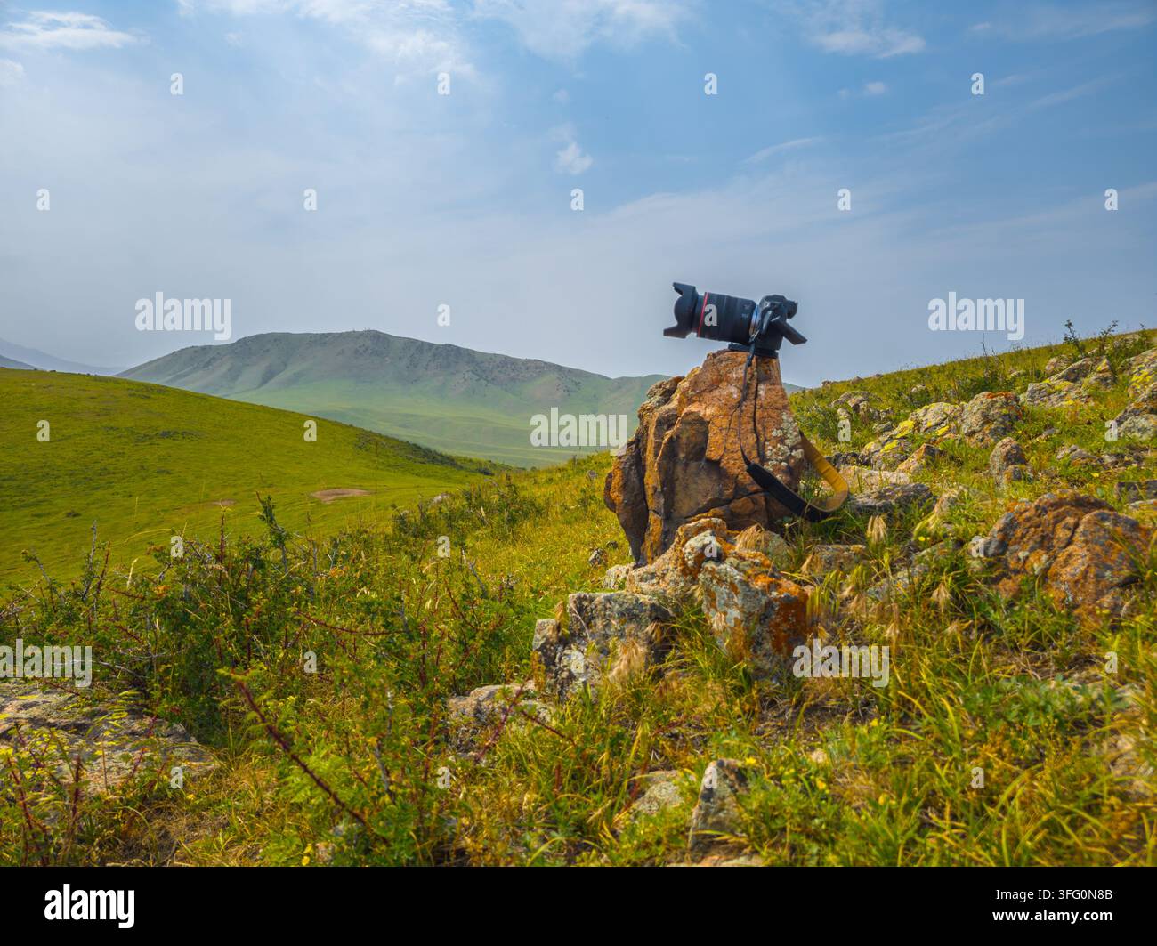 Un appareil photo reflex avec un téléobjectif est monté sur un trépied sur un rocher, surplombant un vaste paysage vallonné au Kirghizistan. La scène capture l'essence de la photographie de paysage sous un ciel partiellement nuageux. Banque D'Images