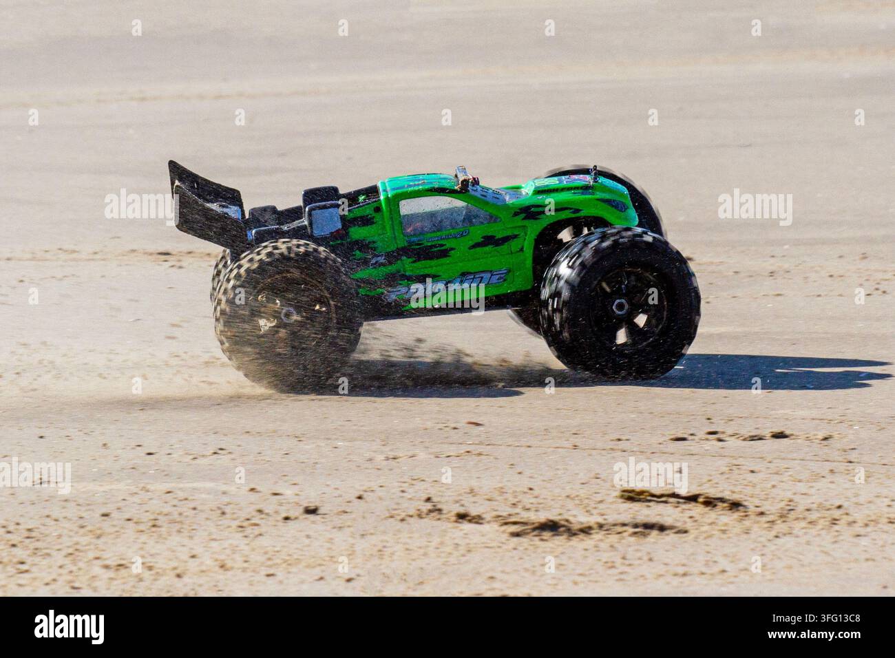 Un camion monstre télécommandé (RC) vert à grande vitesse soulève du sable en traversant une large plage de sable par une journée lumineuse et ensoleillée. Southport, Royaume-Uni. Banque D'Images