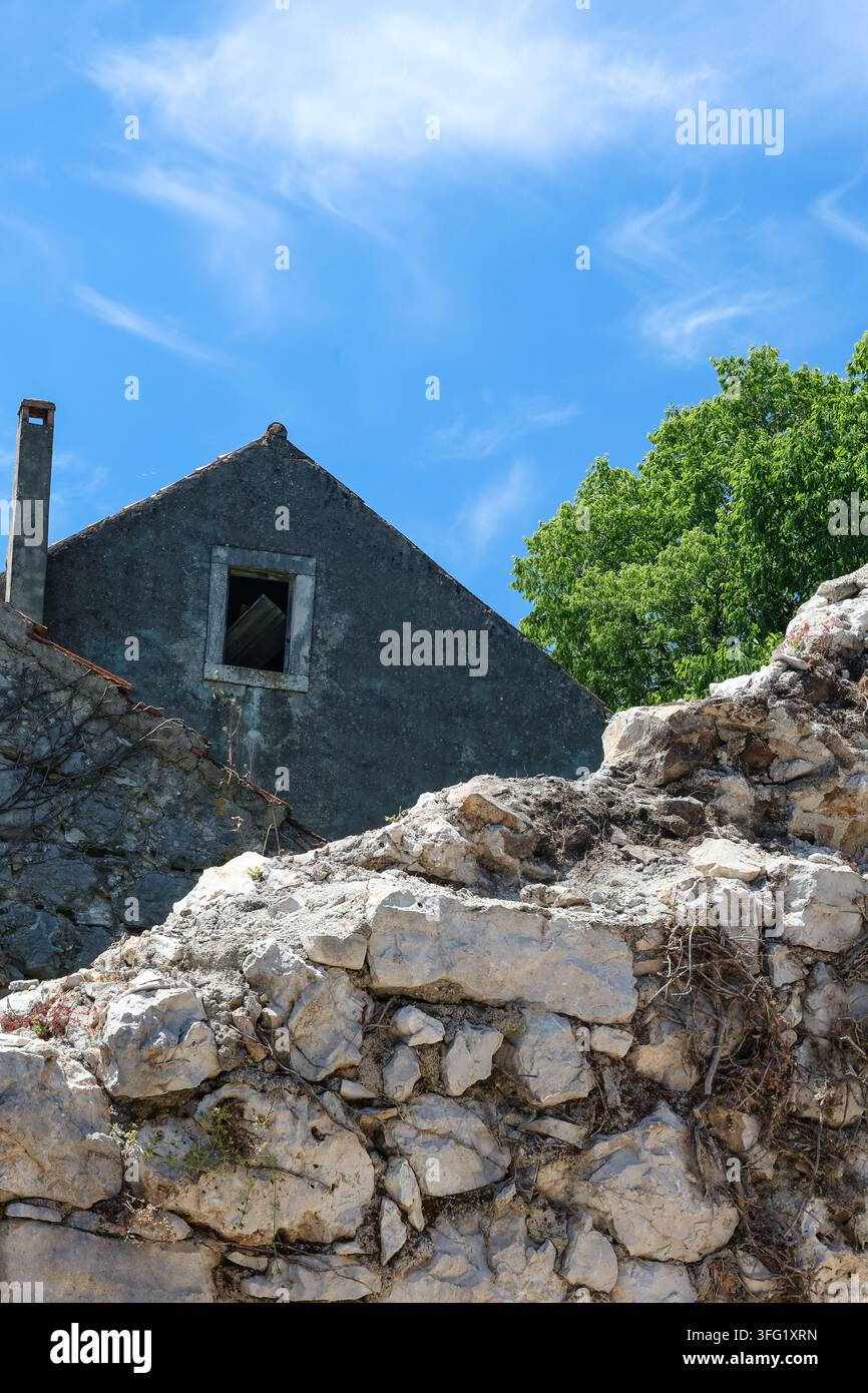 Une maison en pierre abandonnée et un mur de roche en ruine se dressent sous un ciel bleu vif et des nuages filamenteux à côté d'un arbre vert feuillu, évoquant une décadence rustique, soli Banque D'Images