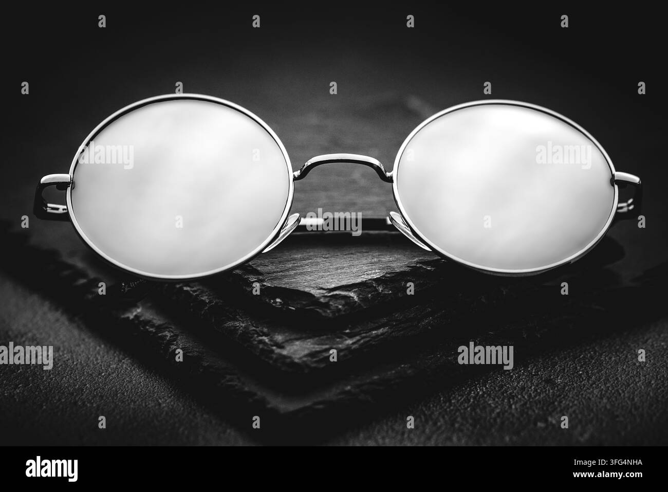 Gros plan en noir et blanc à contraste élevé de lunettes rondes à monture métallique reposant sur un fond d'ardoise sombre, symbolisant la vision ou l'intellect. Banque D'Images