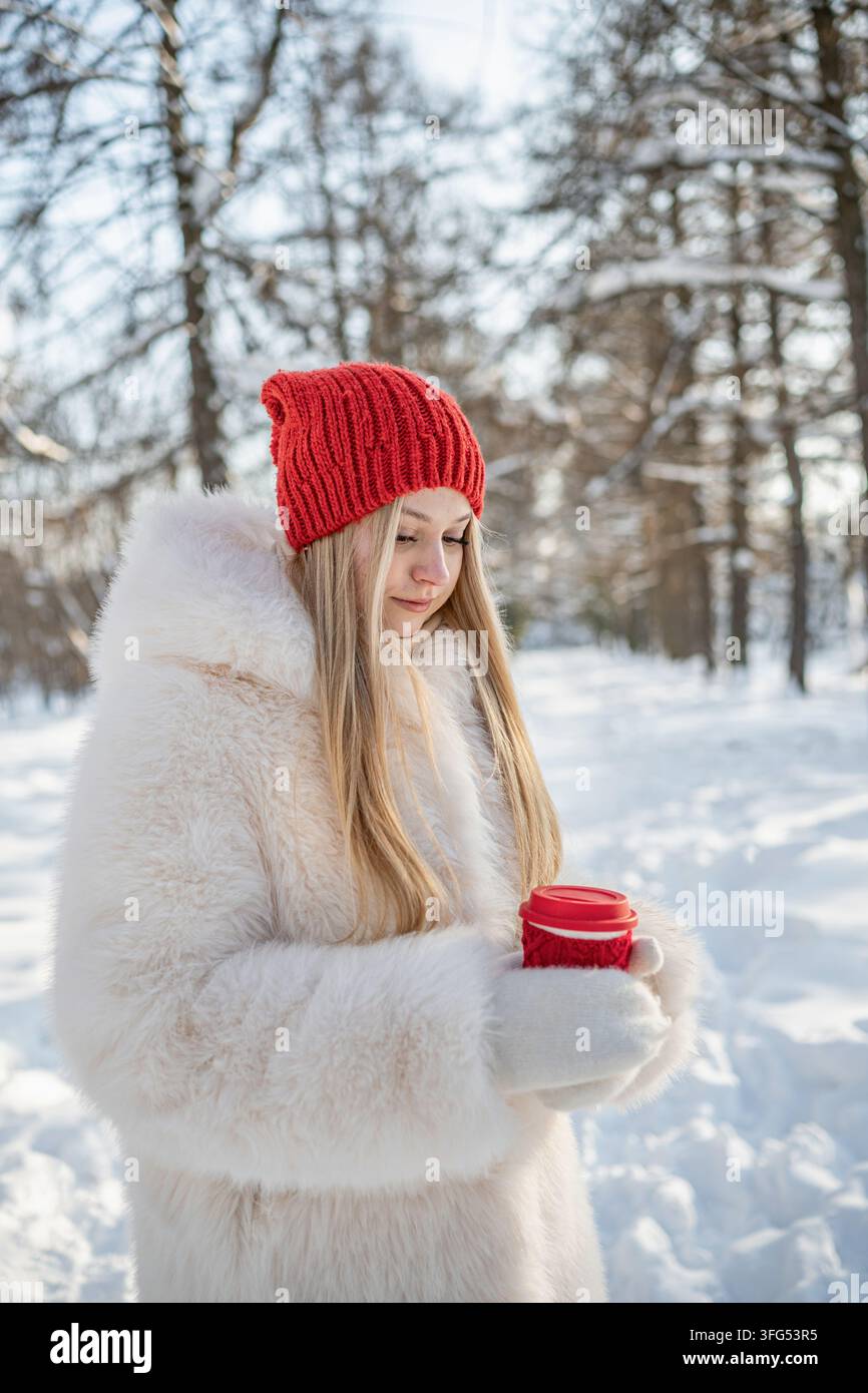 Jeune femme tient une boisson chaude à emporter avec les deux mains dans un parc enneigé, se réchauffant lors d'une promenade hivernale froide et profitant d'une pause saisonnière confortable. Verti Banque D'Images