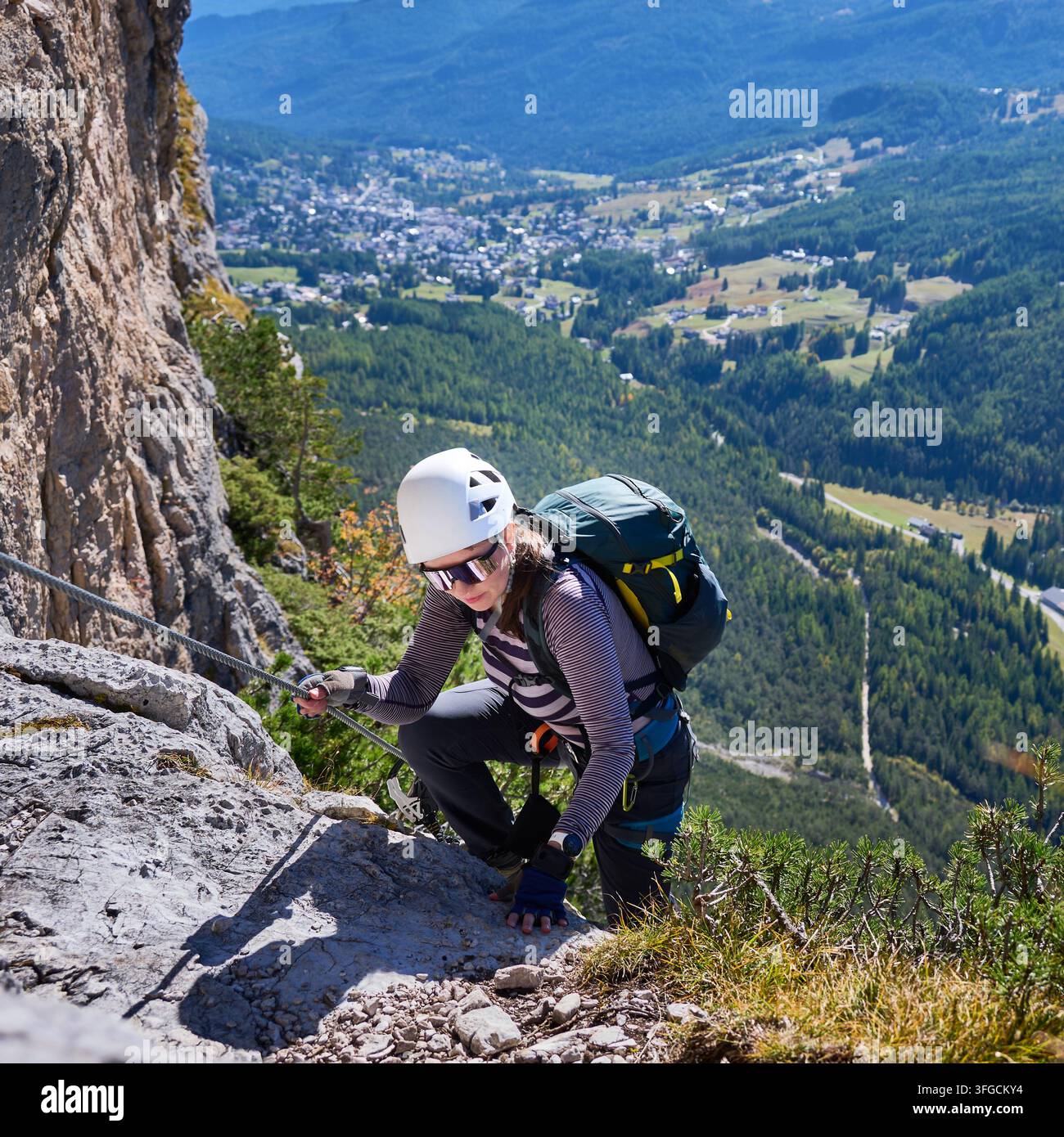Alpiniste féminine gravit une voie ferrata rocheuse tout en s'agrippant à un câble de sécurité en acier au-dessus d'une vallée boisée dans les Dolomites, en Italie. L'image évoque le courage, la concentration et l'aventure alpine active Banque D'Images