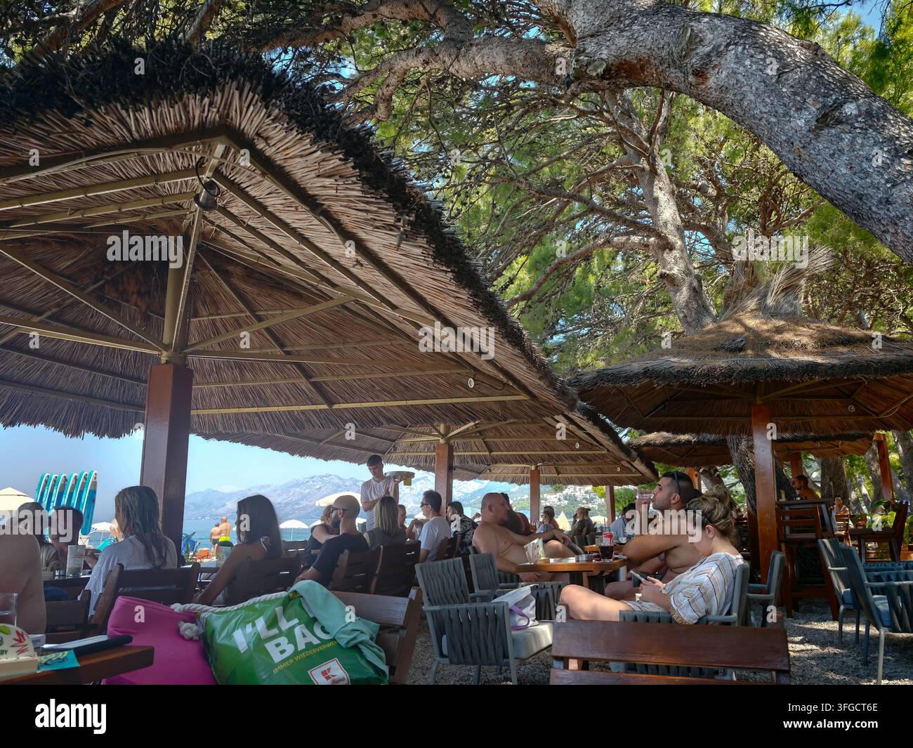 Touristes profitant de boissons rafraîchissantes et de détente sous des parasols en paille ombragés dans un bar de plage animé surplombant la mer étincelante. - Image de stock capturée avec un smartphone