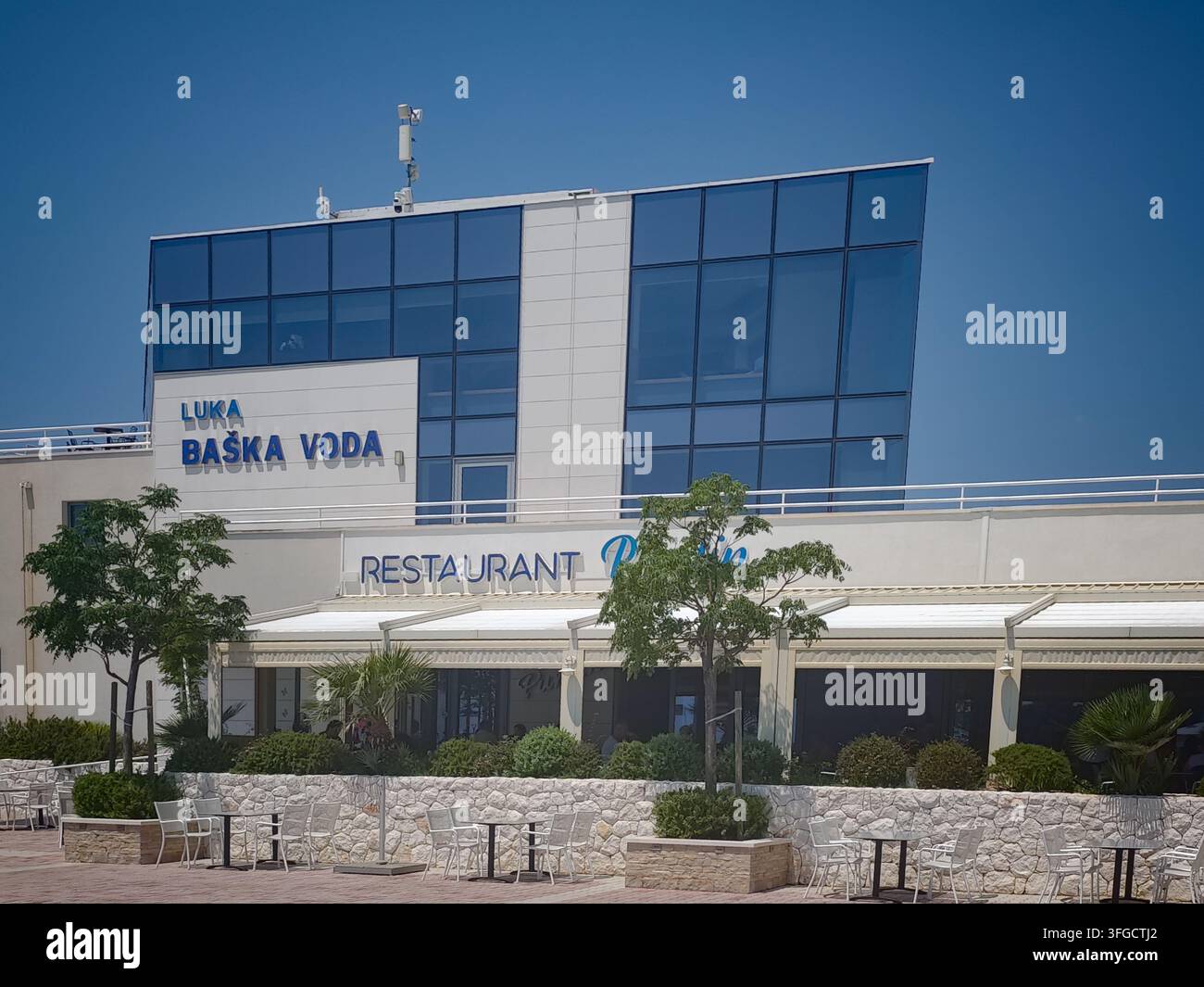 Un bâtiment portuaire contemporain et un restaurant à Baska Voda présentent des fenêtres en verre bleu élégantes et des places assises extérieures sous un ciel bleu éclatant. - Image de stock capturée avec un smartphone
